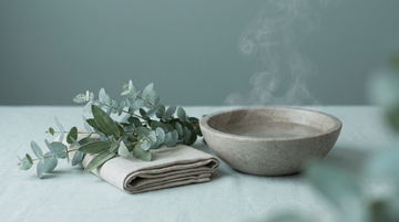 Eucalyptus sprigs and a folded warm compress cloth beside a stone bowl of steaming water, in soft ErasaVir teal light